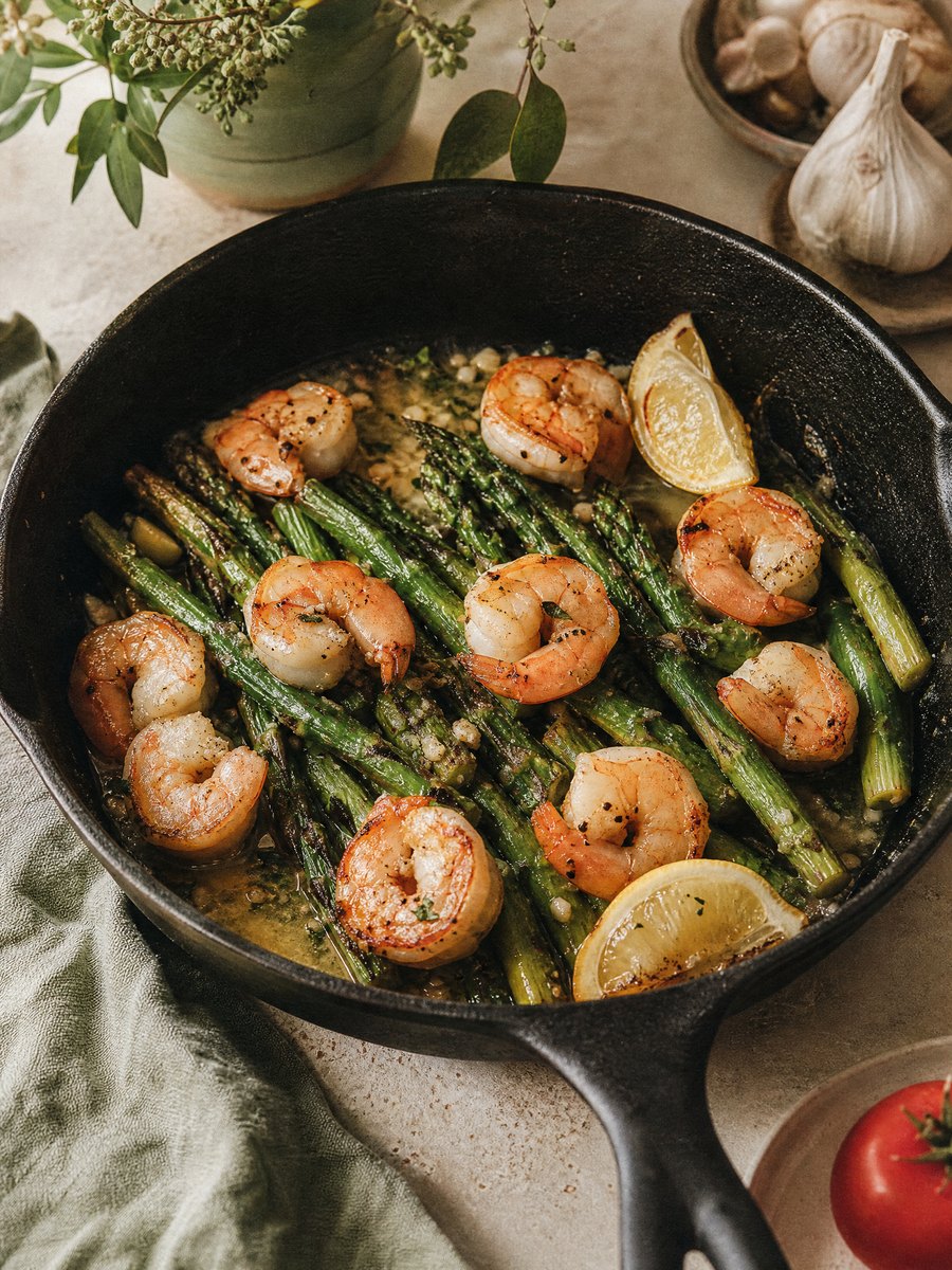 A cast iron skillet filled with seared pink shrimp and bright green asparagus spears, coated in a light garlic butter sauce and garnished with lemon wedges.
